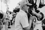 An older man wearing a cap and holding a megaphone stands in a crowd. The megaphone is covered with political stickers, including ones that read 'Trump Won' and 'Women for Trump'. The background is filled with people holding flags, indicating a gathering or protest.