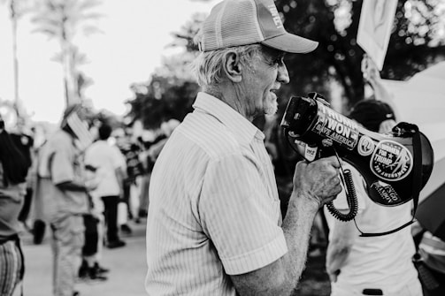 An older man wearing a cap and holding a megaphone stands in a crowd. The megaphone is covered with political stickers, including ones that read 'Trump Won' and 'Women for Trump'. The background is filled with people holding flags, indicating a gathering or protest.