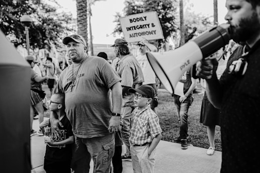 A group of people are gathered outdoors. A man with a beard is holding a megaphone, while another man wearing a cap and a shirt with 'THE TEAM' on it stands next to two children. One child is wearing glasses and the other has a cap. In the background, people are holding signs, one of which reads 'BODILY INTEGRITY & AUTONOMY.' The scene appears to be a protest or gathering.