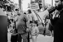 A group of people are gathered outdoors. A man with a beard is holding a megaphone, while another man wearing a cap and a shirt with 'THE TEAM' on it stands next to two children. One child is wearing glasses and the other has a cap. In the background, people are holding signs, one of which reads 'BODILY INTEGRITY & AUTONOMY.' The scene appears to be a protest or gathering.