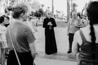 A group of people stand in an outdoor setting, with a priest in a cassock at the center holding what appears to be a rosary or beads. They are surrounded by individuals holding signs, possibly related to a religious or social gathering. The background shows palm trees and a paved pathway, suggesting a park or open public space.