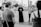 A group of people stand in an outdoor setting, with a priest in a cassock at the center holding what appears to be a rosary or beads. They are surrounded by individuals holding signs, possibly related to a religious or social gathering. The background shows palm trees and a paved pathway, suggesting a park or open public space.