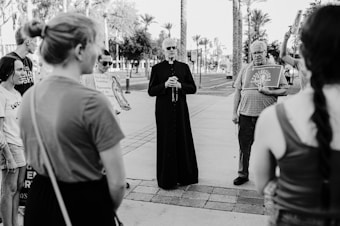 A group of people stand in an outdoor setting, with a priest in a cassock at the center holding what appears to be a rosary or beads. They are surrounded by individuals holding signs, possibly related to a religious or social gathering. The background shows palm trees and a paved pathway, suggesting a park or open public space.