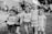 Five young children are standing together, smiling and posing for the camera. They appear to be in a park or an outdoor setting, with some people in the background. The children are wearing casual clothes, with some of their shirts having printed slogans.