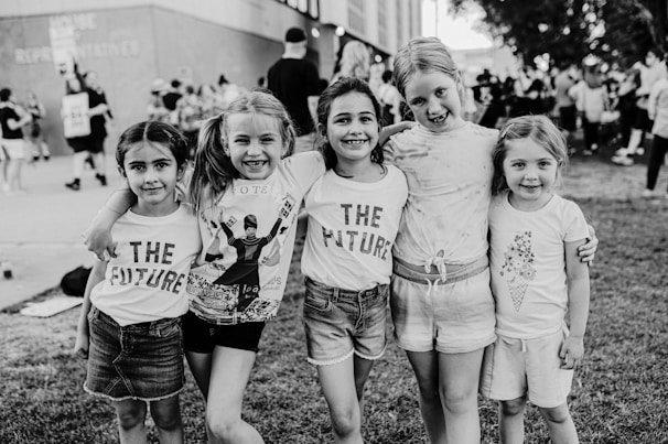 Five young children are standing together, smiling and posing for the camera. They appear to be in a park or an outdoor setting, with some people in the background. The children are wearing casual clothes, with some of their shirts having printed slogans.