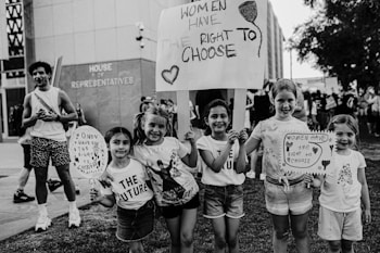 A group of five young girls standing outdoors holding signs that read 'Women have the right to choose'. Three of the girls are holding large posters while two more girls hold smaller ones. There are other people visible in the background. They are standing on grass near a building labeled 'House of Representatives'. Everyone looks happy and full of energy.