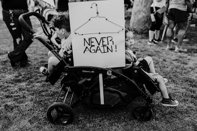 Two children are seated in a double stroller on a grassy area. Attached to the stroller is a sign depicting a wire hanger with the words 'NEVER AGAIN!' written below it. Several people are standing or walking in the background.