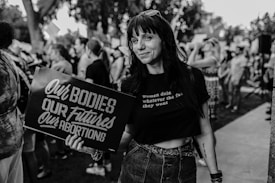A woman holds a sign that reads 'Our Bodies, Our Futures, Our Abortions' at an outdoor protest or rally. She is wearing sunglasses on her head, a black t-shirt with a slogan, and a denim skirt. Other people are gathered in the background, indicating a group event.