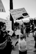 A protest scene with a young child enthusiastically holding a sign that reads 'Girls Should Have Control of Their Own Body.' The child is wearing a tank top and appears determined. Other adults and children are visible in the background, also holding protest signs with various messages. The setting is outdoors, possibly on a street or sidewalk.