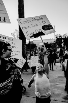 A protest scene with a young child enthusiastically holding a sign that reads 'Girls Should Have Control of Their Own Body.' The child is wearing a tank top and appears determined. Other adults and children are visible in the background, also holding protest signs with various messages. The setting is outdoors, possibly on a street or sidewalk.