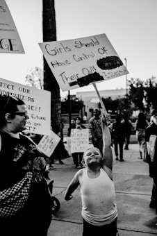 A protest scene with a young child enthusiastically holding a sign that reads 'Girls Should Have Control of Their Own Body.' The child is wearing a tank top and appears determined. Other adults and children are visible in the background, also holding protest signs with various messages. The setting is outdoors, possibly on a street or sidewalk.