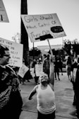 A black and white image captures a protest scene where a child in a tank top energetically holds up a sign reading 'Girls should have control of their own bodies'. Several people in the background are also holding signs and participating in the demonstration.