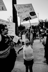 A black and white photograph of a protest or rally, featuring a young child energetically holding up a sign that reads 'Girls Should Have Control of Their Own Bodies.' The child is wearing casual clothing and is surrounded by other protesters, some holding signs. The street setting suggests an urban environment, with blurred figures and trees in the background.
