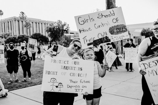 A black and white protest scene with individuals holding signs about women's rights. One sign reads, 'Girls should have control of their own bodies,' held by a young girl. Another sign says, 'Protect our daughters right of choice!' with a drawing of two stick figures.