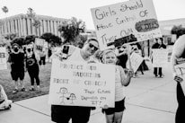 A black and white photo captures a group of people participating in a protest or demonstration. Two individuals in the foreground, an adult and a child, hold signs with messages advocating for women's rights and bodily autonomy. The background shows other participants holding similar protest signs, with trees and a building visible.