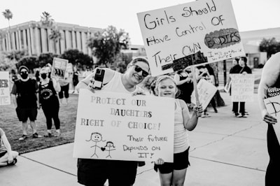 A black and white photo captures a group of people participating in a protest or demonstration. Two individuals in the foreground, an adult and a child, hold signs with messages advocating for women's rights and bodily autonomy. The background shows other participants holding similar protest signs, with trees and a building visible.