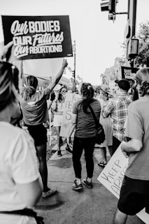 A group of people gathered in protest, holding signs with messages supporting reproductive rights and opposing abortion bans. The setting appears to be a city street. The image is in black and white, capturing several individuals from behind and partially revealing their face expressions.