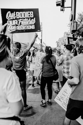 A group of people gathered in protest, holding signs with messages supporting reproductive rights and opposing abortion bans. The setting appears to be a city street. The image is in black and white, capturing several individuals from behind and partially revealing their face expressions.