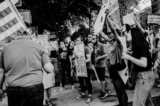 A group of people are gathered in a public space, holding various signs and banners that suggest they are participating in a protest or demonstration. Many individuals appear engaged and animated, expressing their messages strongly. The setting seems to be outdoors during daytime.