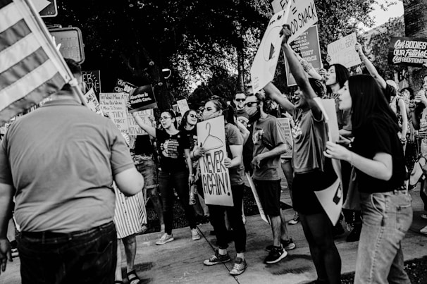 A group of people are gathered in a public space, holding various signs and banners that suggest they are participating in a protest or demonstration. Many individuals appear engaged and animated, expressing their messages strongly. The setting seems to be outdoors during daytime.