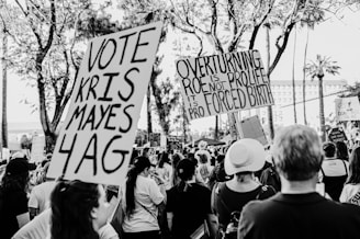 A black and white photograph depicting a crowd of people participating in a protest. They are holding various signs, including ones that read 'Vote Kris Mayes 4 AG' and 'Overturning Roe is not pro life, it is pro forced birth.' The scene is set outdoors with trees in the background, indicating daytime.