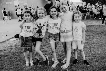 A group of five children stands together on a grassy area, smiling and holding each other in a friendly pose. Three of the children wear shirts with the words 'THE FUTURE,' suggesting a message of optimism and aspiration. The background features a crowd of people, indicating a public or community event.