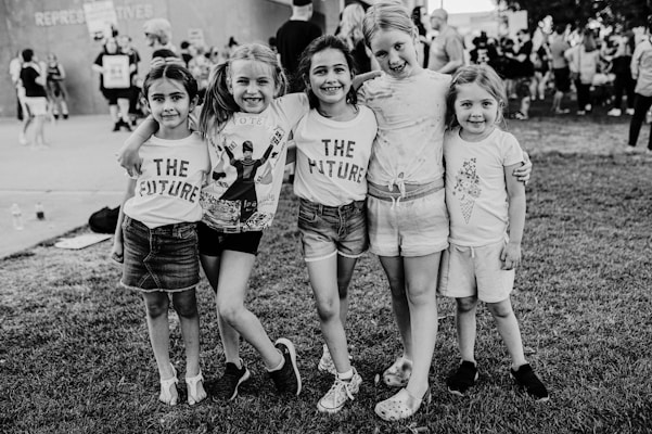 A group of five children stands together on a grassy area, smiling and holding each other in a friendly pose. Three of the children wear shirts with the words 'THE FUTURE,' suggesting a message of optimism and aspiration. The background features a crowd of people, indicating a public or community event.