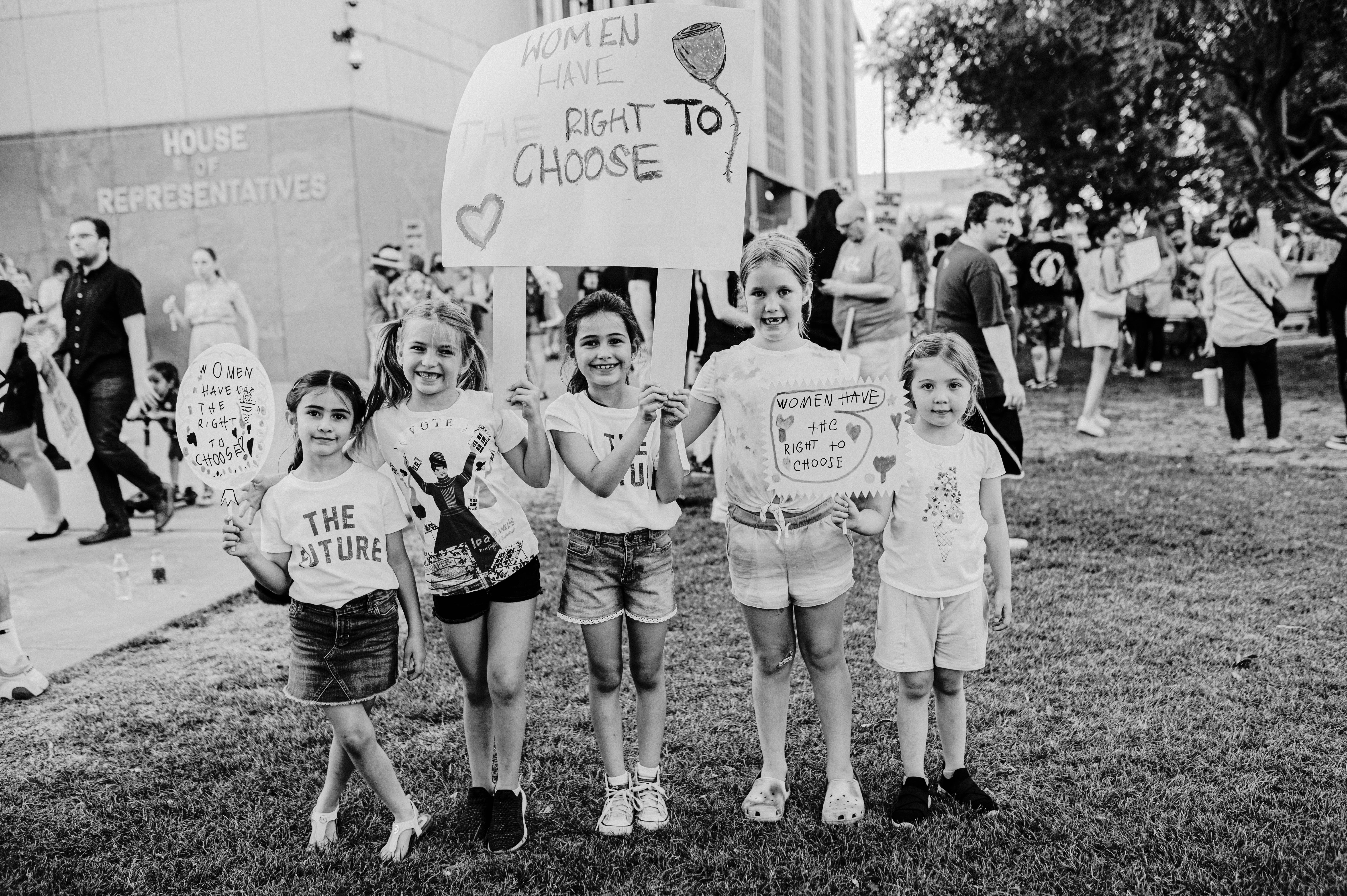 A group of young girls is standing on a grassy area holding protest signs advocating for women's rights. The text on the signs includes messages about women's right to choose. The girls are smiling and appear to be part of a larger gathering, as other people are visible in the background near a building labeled 'House of Representatives'. The image is in black and white.