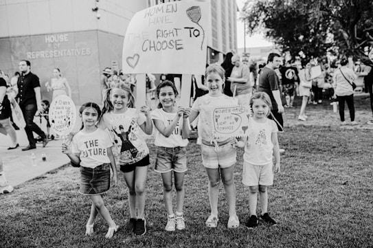 A group of young girls is standing on a grassy area holding protest signs advocating for women's rights. The text on the signs includes messages about women's right to choose. The girls are smiling and appear to be part of a larger gathering, as other people are visible in the background near a building labeled 'House of Representatives'. The image is in black and white.