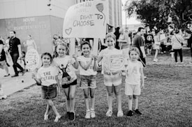 A group of young girls is standing on a grassy area holding protest signs advocating for women's rights. The text on the signs includes messages about women's right to choose. The girls are smiling and appear to be part of a larger gathering, as other people are visible in the background near a building labeled 'House of Representatives'. The image is in black and white.