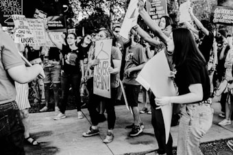 A group of people is gathered outdoors, holding up signs and banners with messages related to abortion rights and healthcare. The crowd appears animated and engaged, with some individuals shouting or raising their fists. The scene is captured in black and white, adding a dramatic tone to the composition.