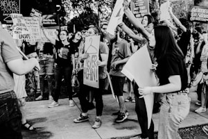A group of people is gathered outdoors, holding up signs and banners with messages related to abortion rights and healthcare. The crowd appears animated and engaged, with some individuals shouting or raising their fists. The scene is captured in black and white, adding a dramatic tone to the composition.