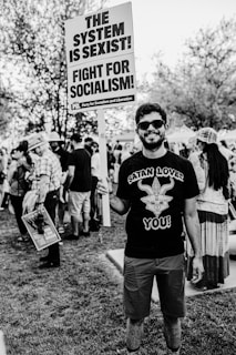 A man stands in a park holding a large protest sign with bold text. The sign reads 'The System is Sexist! Fight for Socialism!' Other protestors can be seen in the background, some holding signs. The man is wearing sunglasses and a t-shirt with a graphic design.