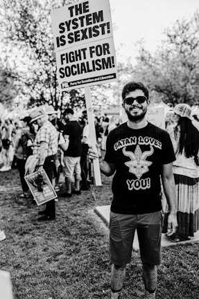 A person stands in a park holding a protest sign that reads 'THE SYSTEM IS SEXIST! FIGHT FOR SOCIALISM!' There are other people in the background, some with hats and some holding signs, suggesting a protest or rally. The person is wearing sunglasses and a shirt with a graphic design.