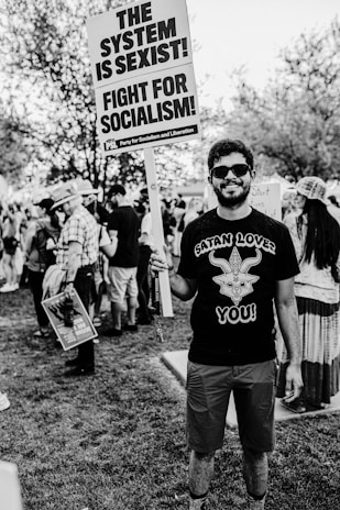 A person stands in a park holding a protest sign that reads 'THE SYSTEM IS SEXIST! FIGHT FOR SOCIALISM!' There are other people in the background, some with hats and some holding signs, suggesting a protest or rally. The person is wearing sunglasses and a shirt with a graphic design.