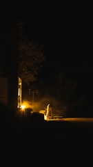 A dimly lit scene showcases a person working near a light source outside a building during nighttime. The environment is shrouded in darkness, with a warm glow emanating from the light, casting long shadows. The activity appears to be work-related, possibly involving construction or maintenance.