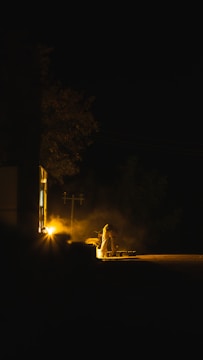 A dimly lit scene showcases a person working near a light source outside a building during nighttime. The environment is shrouded in darkness, with a warm glow emanating from the light, casting long shadows. The activity appears to be work-related, possibly involving construction or maintenance.