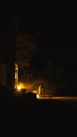 A dimly lit scene showcases a person working near a light source outside a building during nighttime. The environment is shrouded in darkness, with a warm glow emanating from the light, casting long shadows. The activity appears to be work-related, possibly involving construction or maintenance.