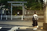 A person in a school uniform stands at a crosswalk, holding a bag and wearing a backpack. In the background, there is a traditional stone torii gate and a stone lantern, suggesting a Japanese setting. Greenery surrounds the gate, and a blurred figure is visible in the distance.