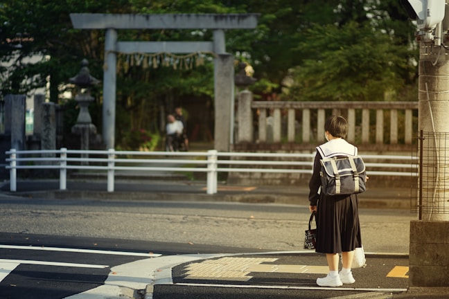 A person in a school uniform stands at a crosswalk, holding a bag and wearing a backpack. In the background, there is a traditional stone torii gate and a stone lantern, suggesting a Japanese setting. Greenery surrounds the gate, and a blurred figure is visible in the distance.