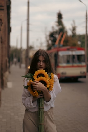A person with long brown hair is standing on a street holding a bouquet of large sunflowers. They are wearing a white long-sleeved blouse and beige pants. In the background, there's a blurred image of a red and white trolleybus and lampposts lining the sidewalk.
