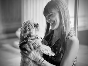 A woman is holding a small dog close to her face, smiling as they share a moment. The image captures a sense of joy and companionship between the two, with the background softly blurred.