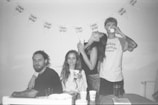 A monochrome photo depicts four people celebrating at an event. Two are seated on a couch, while two are standing. Decorations on the wall include a banner with 'Happy Birthday' text. Various cups are placed on the table.