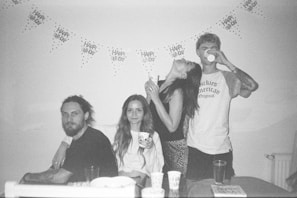 A monochrome photo depicts four people celebrating at an event. Two are seated on a couch, while two are standing. Decorations on the wall include a banner with 'Happy Birthday' text. Various cups are placed on the table.