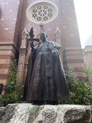 A bronze statue of a religious figure holding a bird is positioned in front of a brick church wall with intricate architectural details. The background features a large circular stained glass window. Greenery surrounds the base of the statue, adding a natural element to the scene.