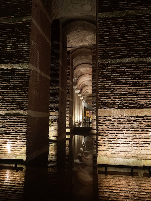 A series of ancient, massive stone columns line a dimly lit underground chamber. The architecture features arches that create a repeating pattern, stretching into the distance. Soft lighting accentuates the texture of the aged bricks, casting reflections in the shallow water on the floor.