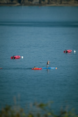 A person is paddleboarding on a calm body of water, accompanied by another person in a red kayak. The water is a clear blue, with gentle ripples. In the background, there are several red kayaks floating near white buoys.