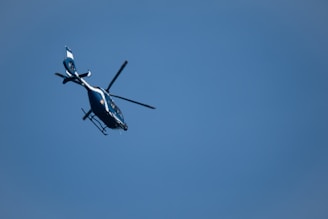A helicopter with a sleek design is flying against a backdrop of a clear blue sky. It appears to be angled slightly upward as it moves through the air.