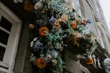 An ornate floral arrangement featuring a variety of flowers, including orange, blue, and white blooms, is displayed against a stone building near a window. The arrangement appears lush and vibrant with greenery interspersed among the flowers.
