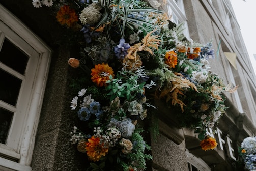 An ornate floral arrangement featuring a variety of flowers, including orange, blue, and white blooms, is displayed against a stone building near a window. The arrangement appears lush and vibrant with greenery interspersed among the flowers.